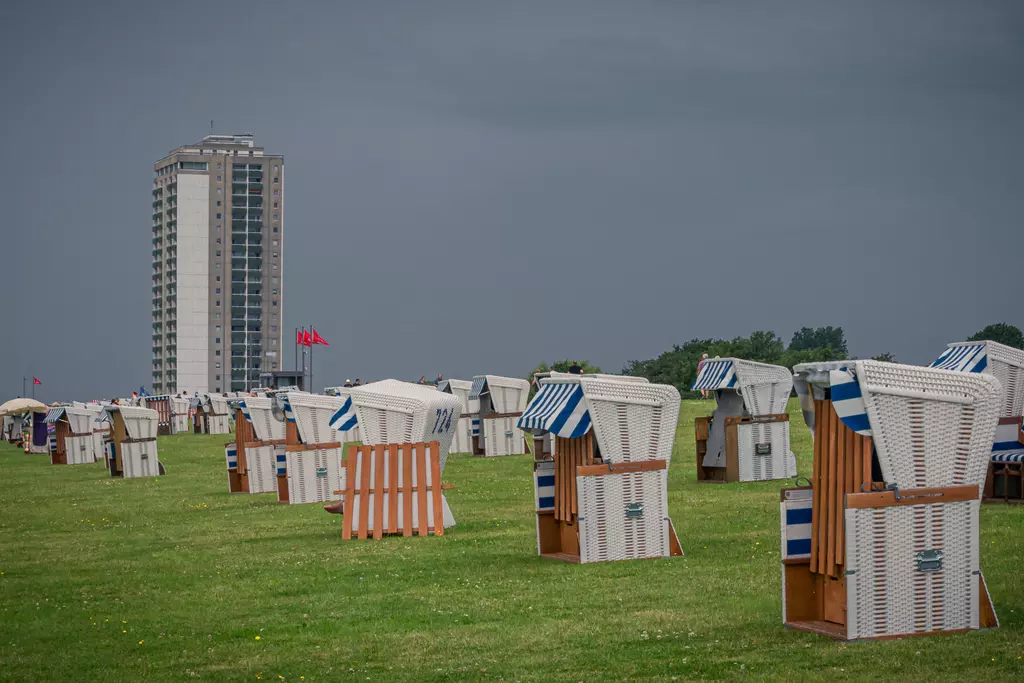 Rows of colorful beach chairs on green grass, with a tall building and dark clouds in the background.