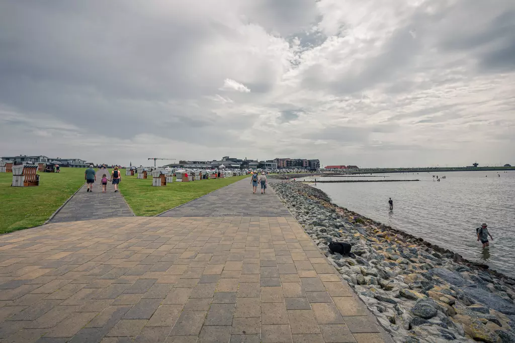 People walk along a paved path by the coast, lined with grass and stones, under a cloudy sky in the background.