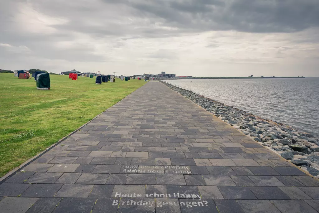 A paved walkway runs along the coast, surrounded by green spaces and beach chairs, under a cloudy sky.