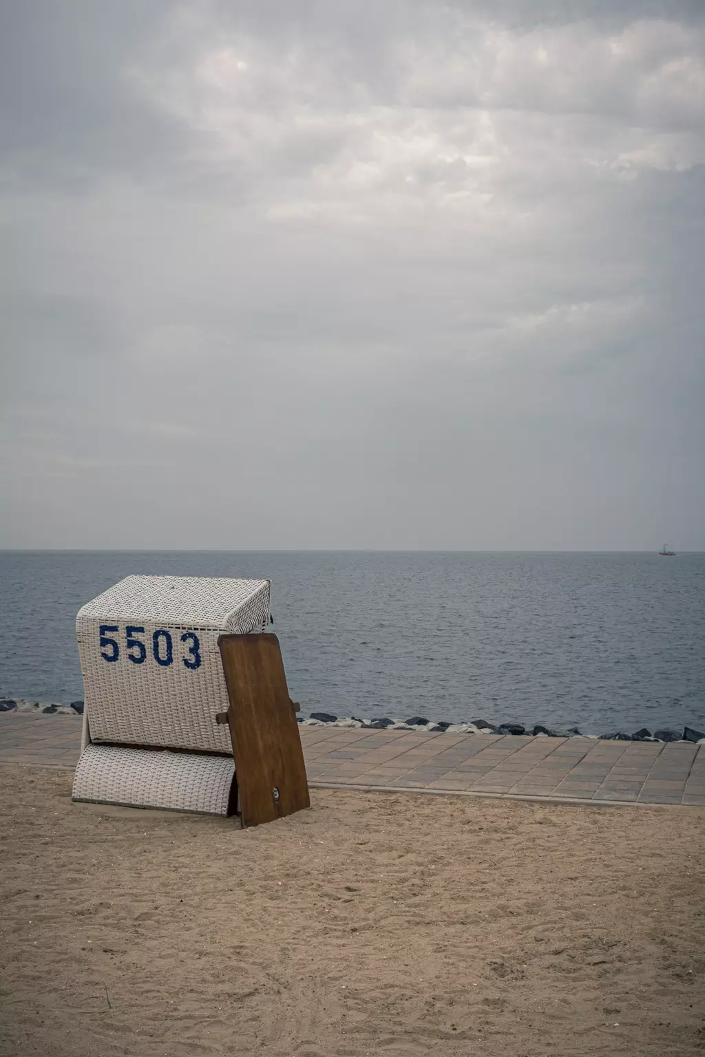 A beach chair numbered 5503 sits on the sand, overlooking calm water with a gray sky in the background.