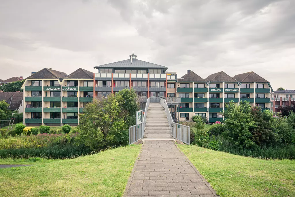 A path leads to a multi-story building with balconies, surrounded by trees and grass. The sky is overcast.