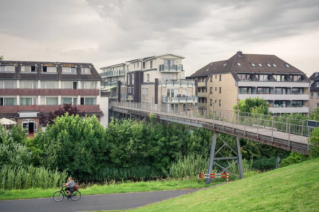 Two modern buildings, a cyclist on a path, a footbridge, surrounded by trees and grass.