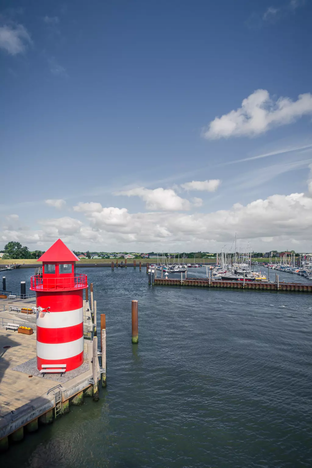 Leuchtturm auf Föhr: Ein roter und weiß gestreifter Leuchtturm steht an einer Hafenmole, umgeben von Booten und ruhigem Wasser.