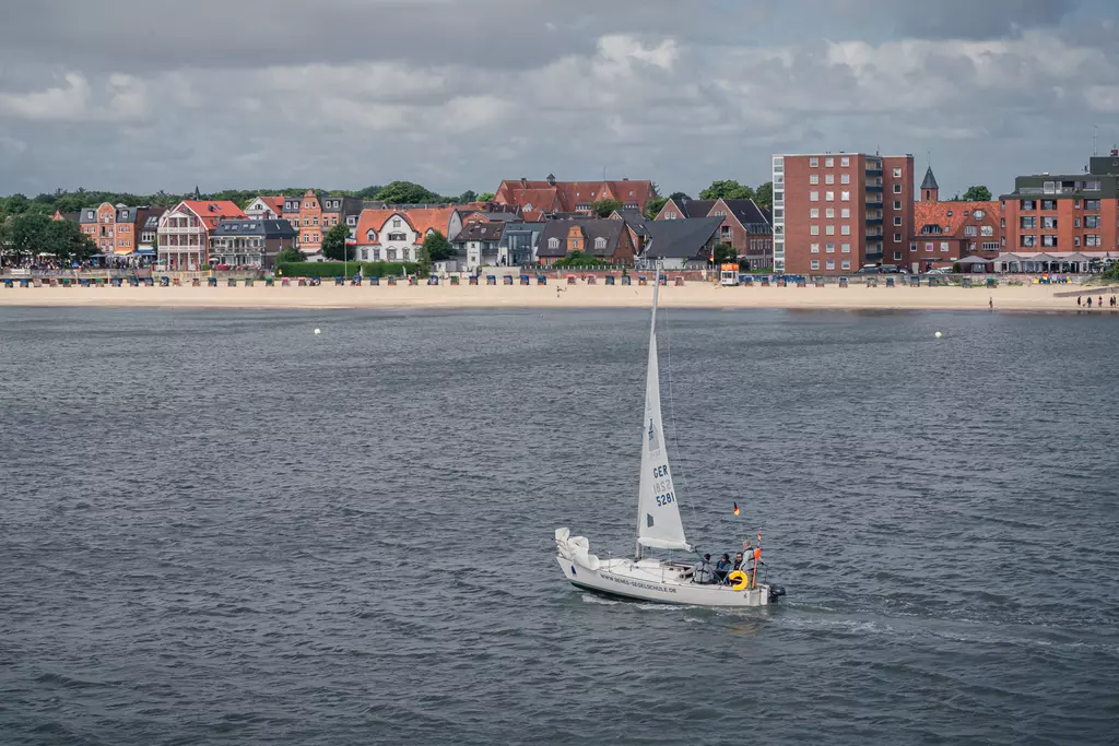 Föhr: Ein Segelboot fährt auf dem Wasser vorbei, während die Küste mit bunten Häusern und einem Sandstrand im Hintergrund zu sehen ist.