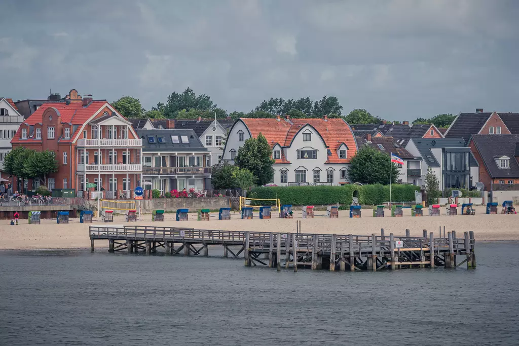 Strand in Föhr: Holzsteg führt zur Küste, mit bunten Strandkörben und historischen Gebäuden im Hintergrund.