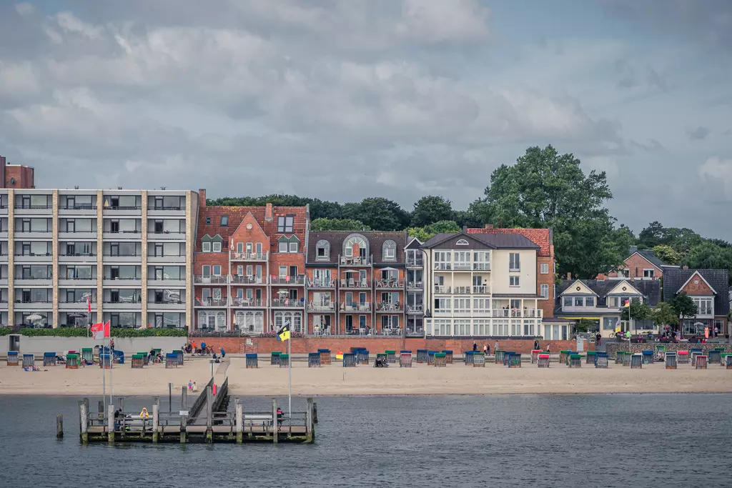 Eine Uferpromenade mit mehreren Gebäuden, bunten Strandkorb-Bereichen und einem steinernen Steg am Wasser unter bewölktem Himmel.