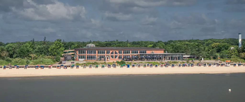 Strand auf Föhr: Ein modernes Gebäude am Sandstrand mit vielen Strandkörben und Wald im Hintergrund.