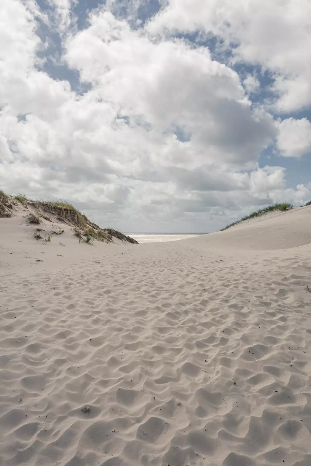 Weicher Sandweg zwischen Dünen unter einem bewölkten Himmel, der zum Meer führt. Grüne Gräser wachsen in den Dünen.