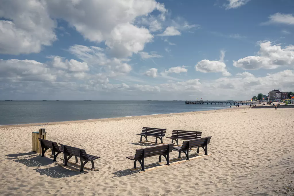 Strand auf Föhr: Mehrere Holzbänke stehen im Sand, Blick auf das ruhige Wasser und einen Pier in der Ferne.