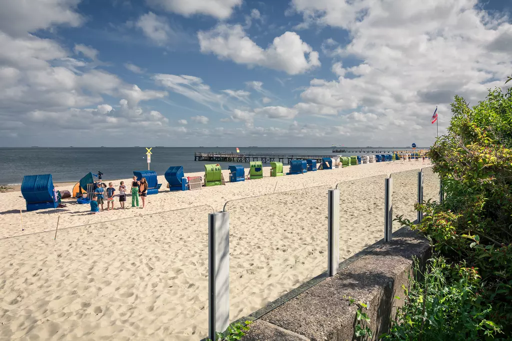 Strand auf Föhr: Sandstrand mit zahlreichen Strandkörben, ein paar Personen stehen am Wasser, Holzpier im Hintergrund.