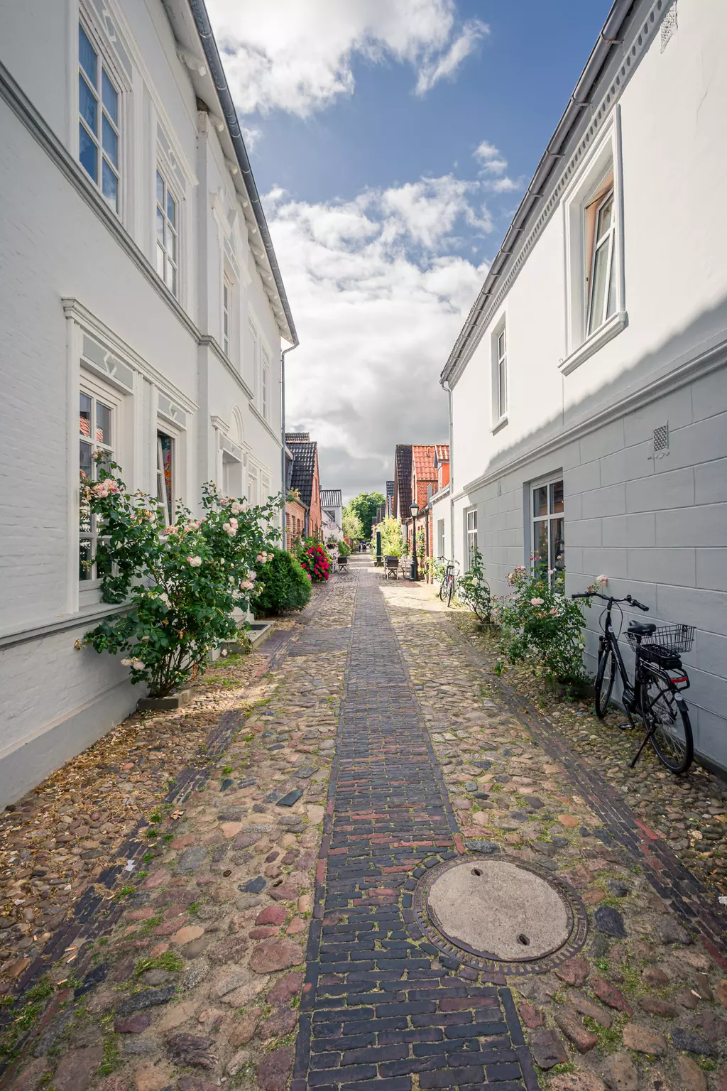 Föhr: Eine gepflasterte Gasse zwischen weißen Gebäuden, gesäumt von grünen Sträuchern und Fahrrädern.