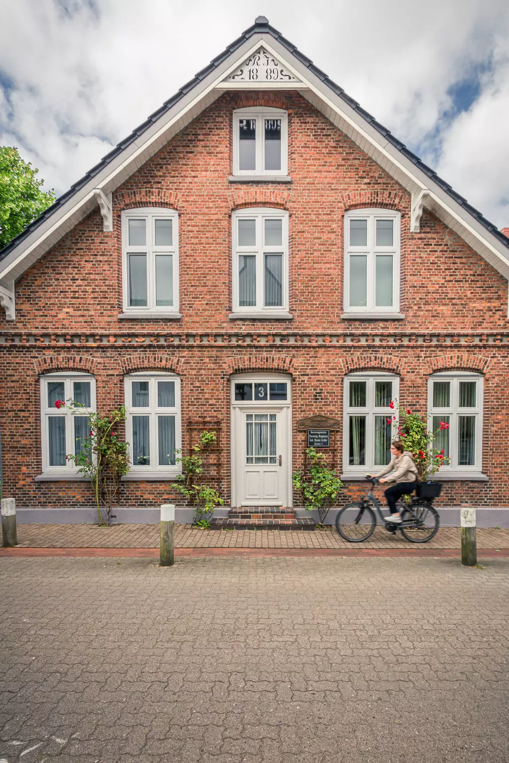 Ein rotes Backsteinhaus mit weißem Eingang, großen Fenstern und Fahrradfahrer auf dem Bürgersteig, Blumen an der Wand.