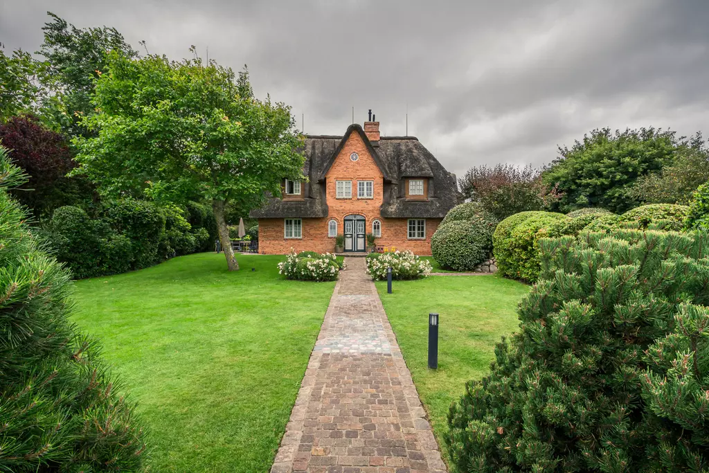 Haus in Sylt: Ein traditionelles, reetgedecktes Haus mit brauner Backsteinfassade, umgeben von gepflegtem Grün und Bäumen.