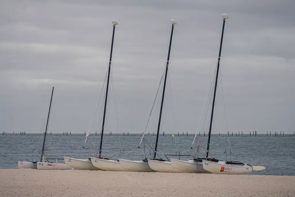 Segelboote auf Sylt: Vier Segelboote stehen am Strand, ihre Masten ragen in den grauen Himmel über dem Wasser.