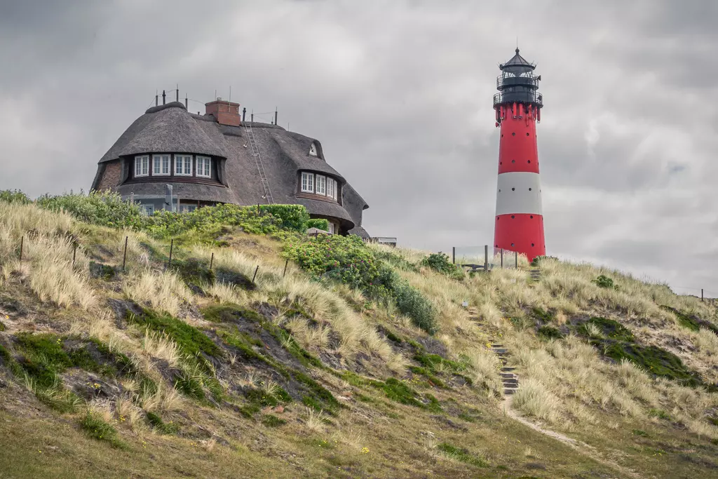 Ein roter Leuchtturm steht neben einem traditionellen, strohgedeckten Haus auf einer Dünenlandschaft mit grünem Gras und Büschen.