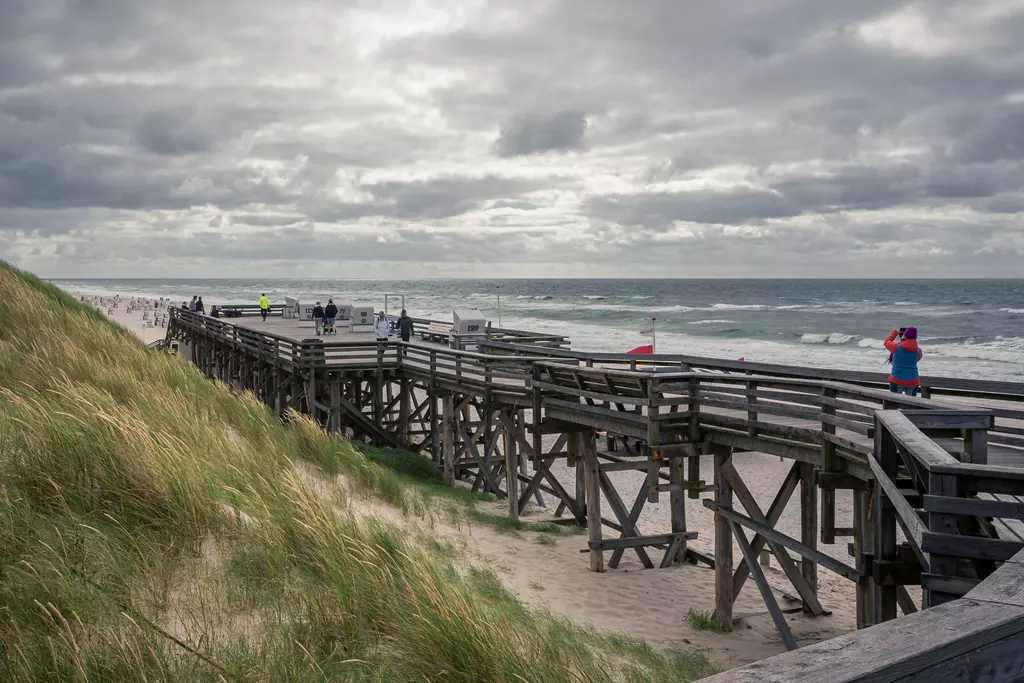 Strandpromenade auf Sylt: Eine hölzerne Brücke führt über den Sand, umgeben von Gräsern und Wolken am Himmel.