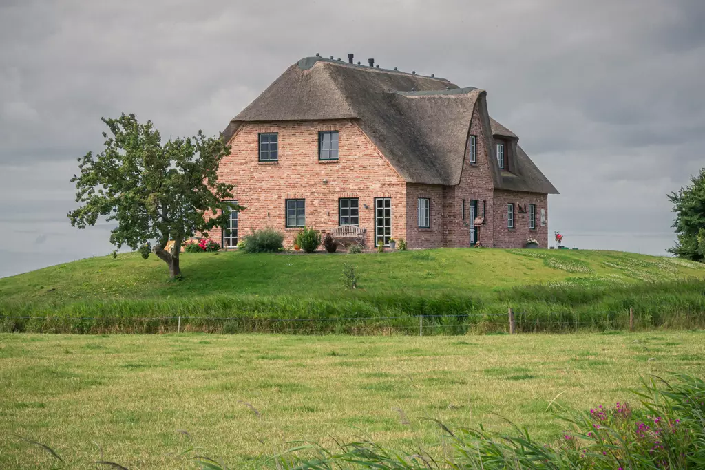 Ein großes, strohgedecktes Haus aus roten Ziegeln auf einem Hügel, umgeben von grünen Wiesen und einigen Bäumen.