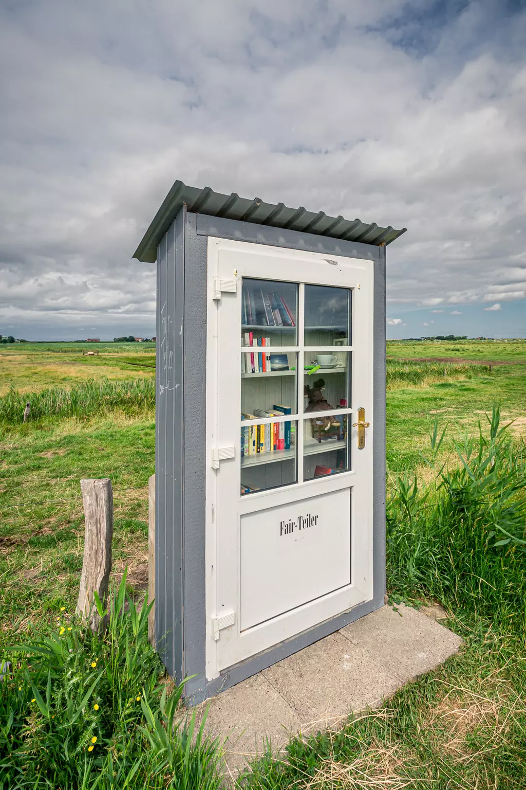 Ein kleiner, grauer Bücherkasten steht in einer grünen Wiese, umgeben von weitem Himmel und einzelnen Wolken.