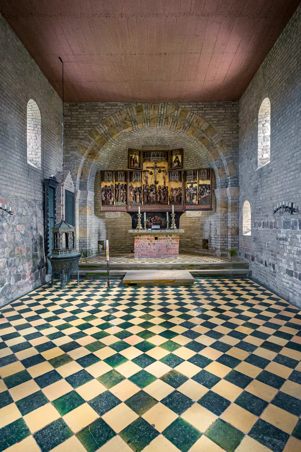 Ein historischer, schlichter Kirchenraum mit einem gemusterten Boden, einem Altar mit einem großen, farbenfrohen Altarbild im Hintergrund.