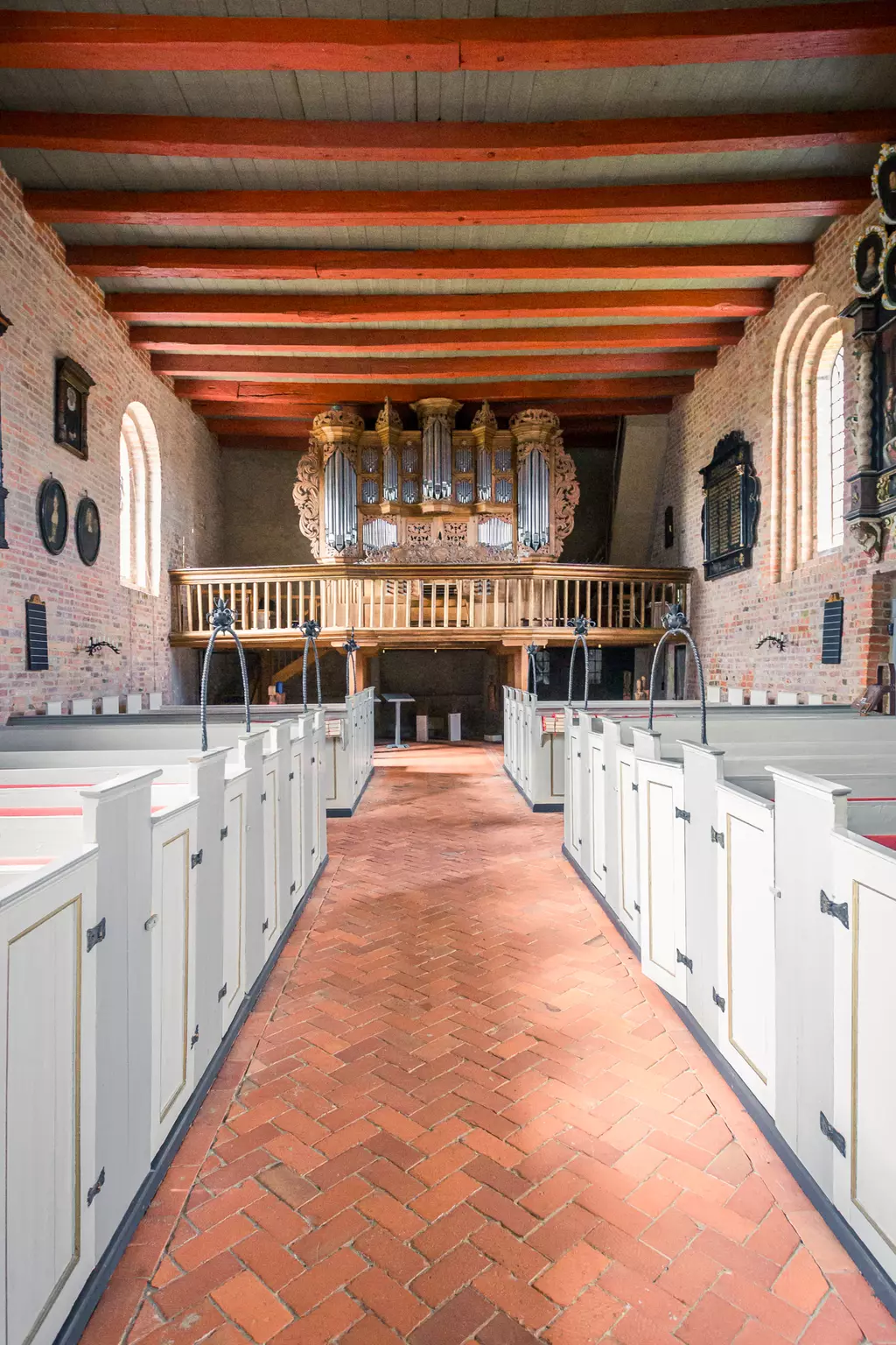 Eine Sicht von innen auf eine Kirche mit einem langen Gang, weißen Bänken und einer kunstvollen Orgel im Hintergrund.