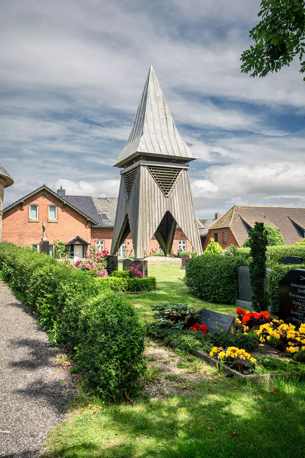 Ein schmaler Holzbau mit einem spitzen Dach steht zwischen Blumenbeeten und Gräbern in einer grünen Umgebung.