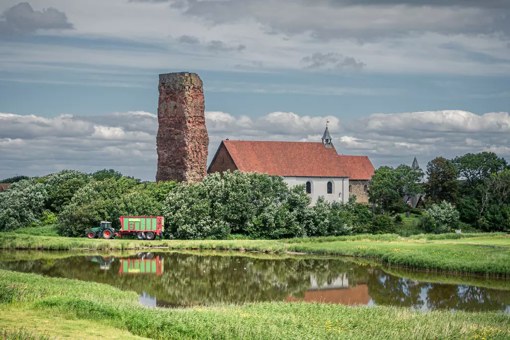 Ein alte Kirche mit rotem Dach, umgeben von Bäumen, neben einem steinernen Turm und einem ruhigen Teich im Vordergrund.