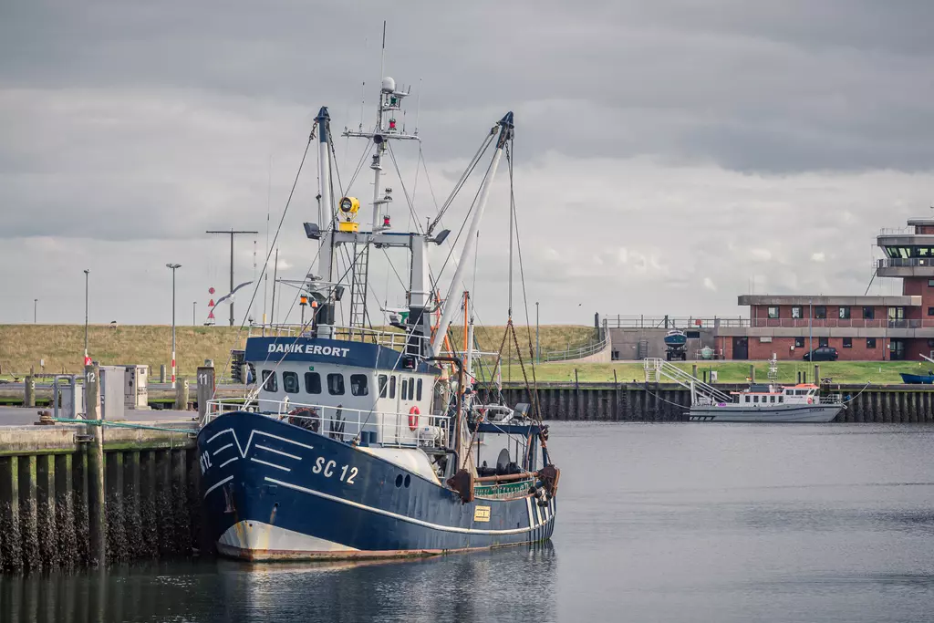 A fishing vessel painted blue is docked in the harbor, surrounded by water and clouds above, with a building in the background.