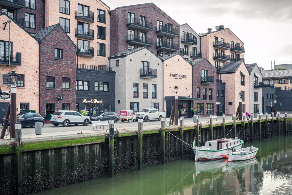 Modern buildings in various colors line the waterfront with boats in the water and cars parked along the street. A serene harbor view.