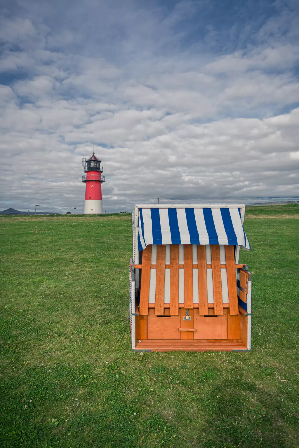 A beach chair with blue and white stripes sits on green grass, with a red lighthouse in the background under a cloudy sky.