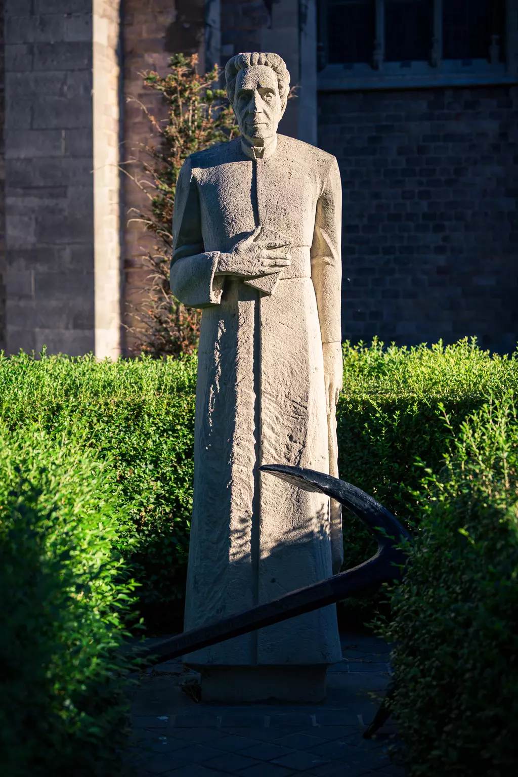 A statue of a figure in a long robe stands among green bushes, with an anchor in the foreground.