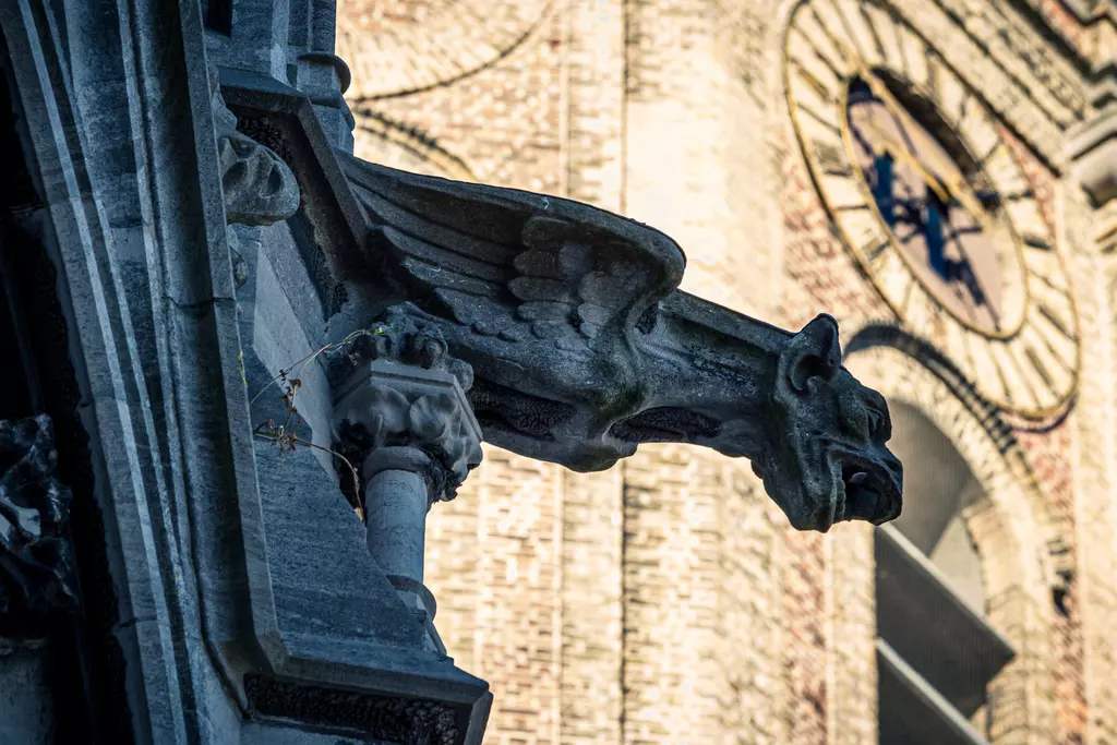 A stone gargoyle with wings and an animal-like face, set against a historic brick wall.