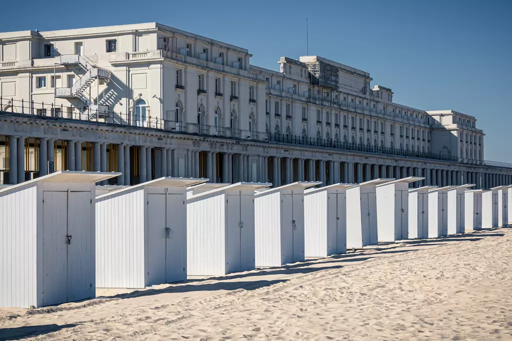 Rows of white beach cabins on light sand, with an elegant historic building in the background under a clear sky.