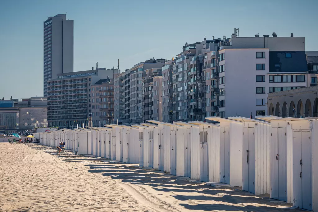 A row of white beach cabins stands in the sand, with modern buildings rising in the background.