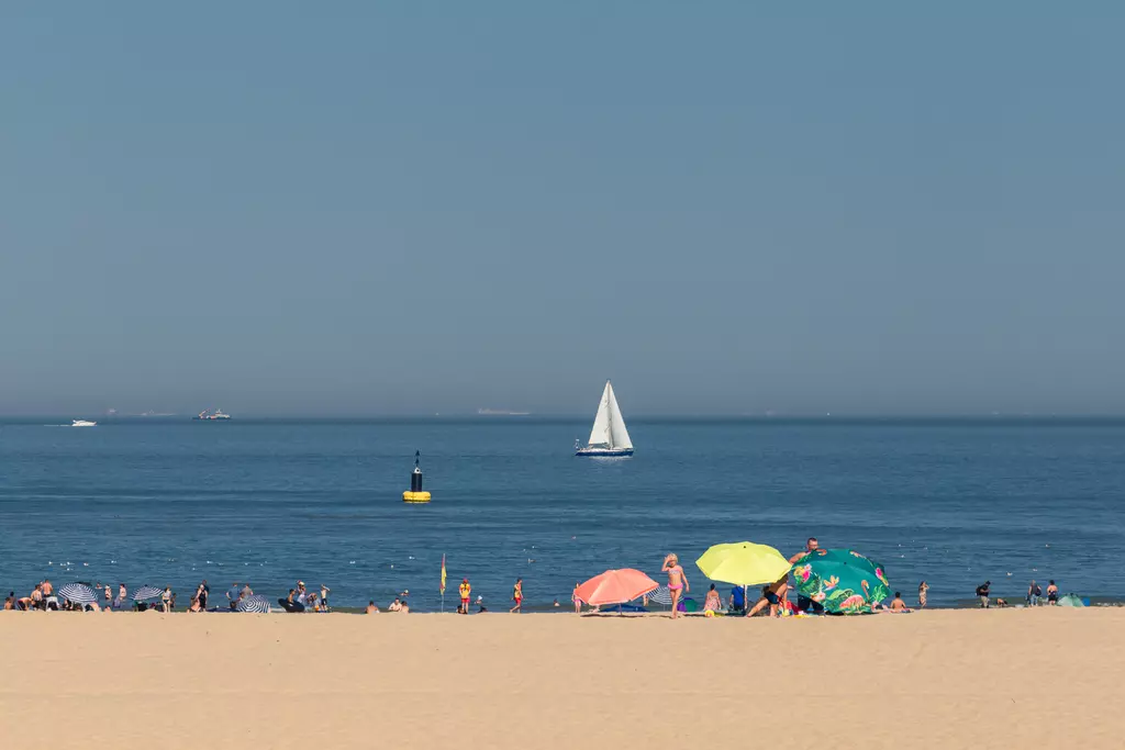 View of a beach with people, colorful umbrellas, and a sailboat on the water. The sky is clear and blue.