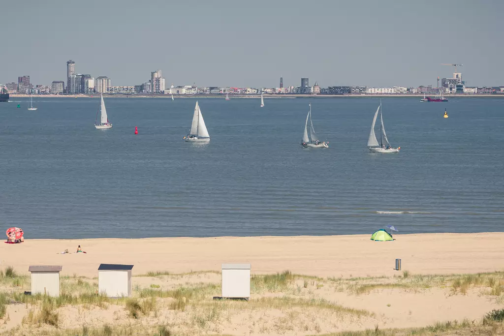 Soft sandy beach with sailboats on the water and a city skyline in the background. Green tent and beach chairs at the shore.