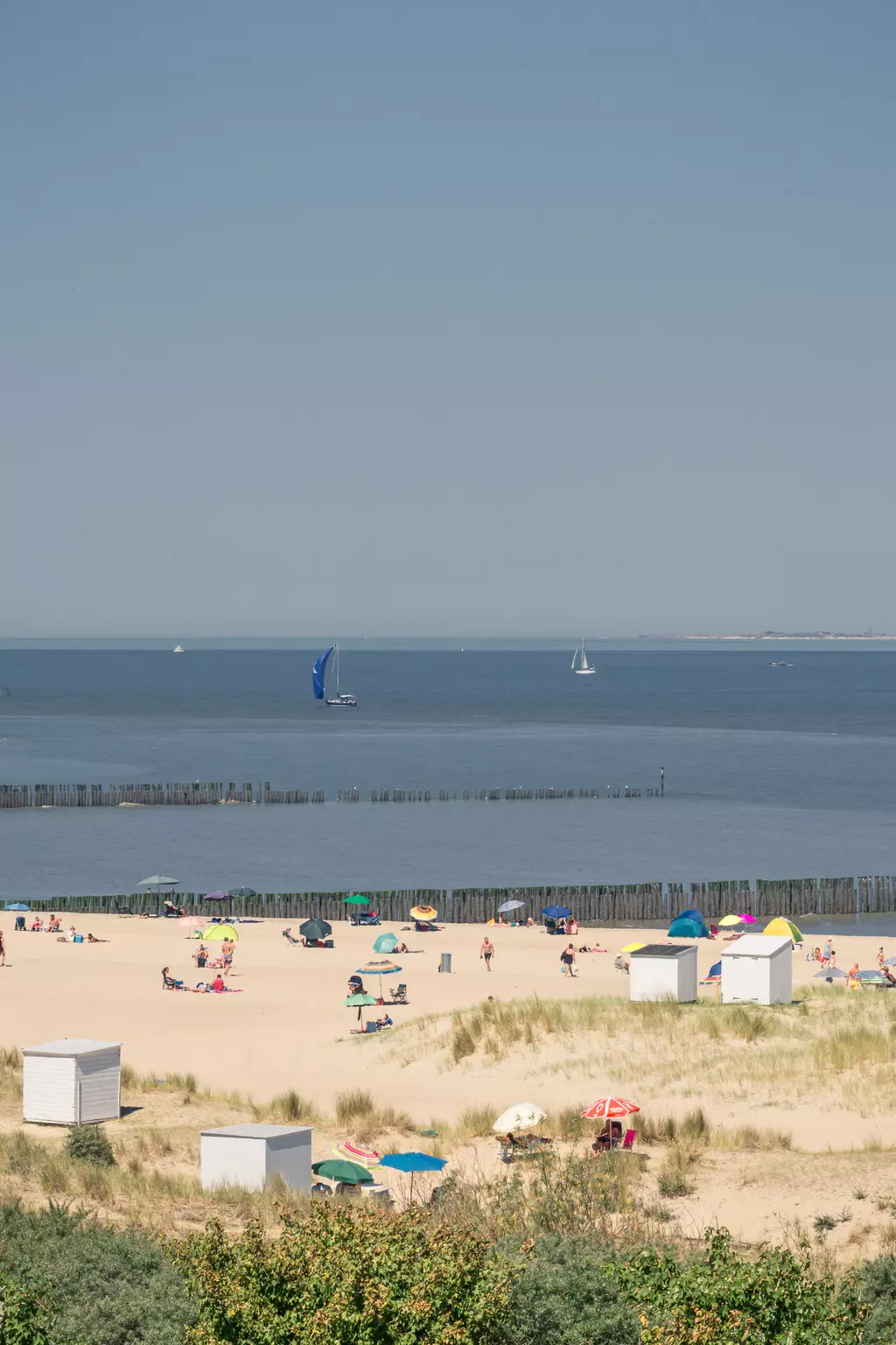 A sunny beach with sun loungers and colorful umbrellas, sailboats visible on the water in the distance.