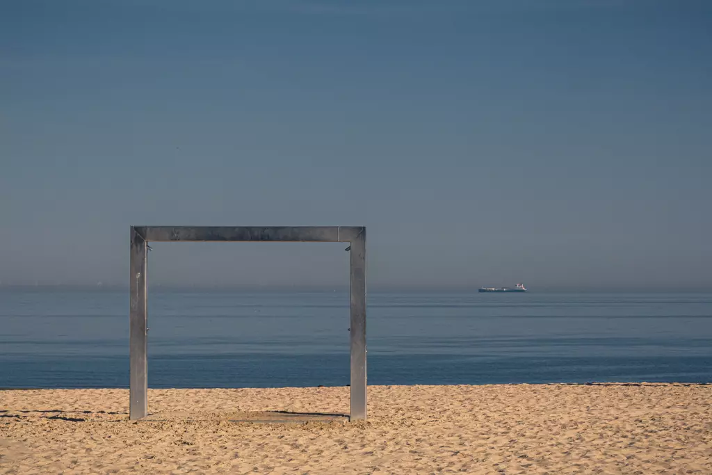 A square metal frame stands in the sand, with calm water and a ship visible on the horizon in the background.