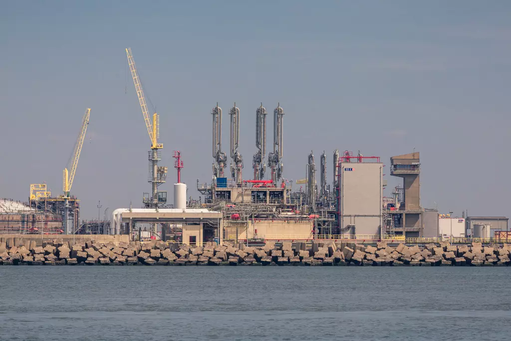 Industrielle Anlage mit hohen Türmen, Rohren und Kränen, die am Ufer stehen, umgeben von Wasser und Felsen.