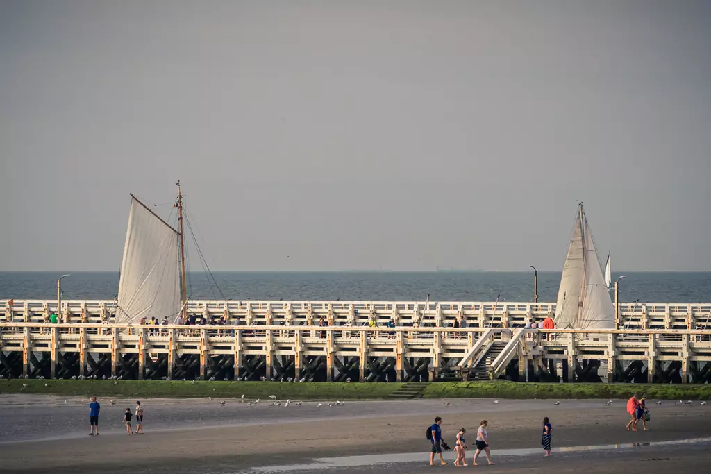 Two sailboats are sailing on the water while people walk on a wooden pier. The sky is cloudy.