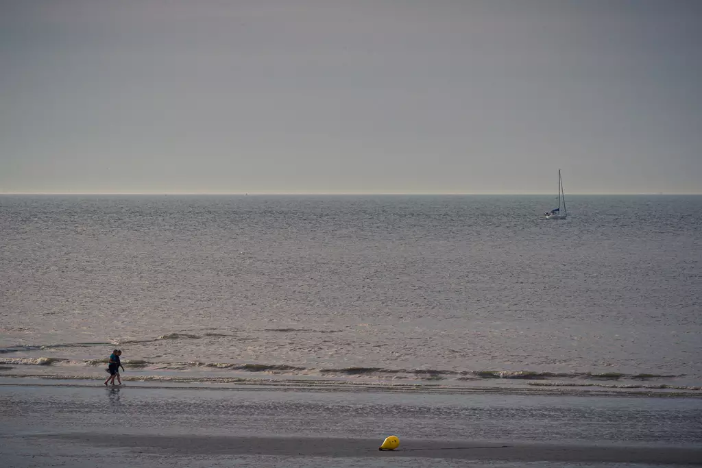 A person walks along the beach as a yellow buoy floats in the water, with a sailboat visible in the distance.