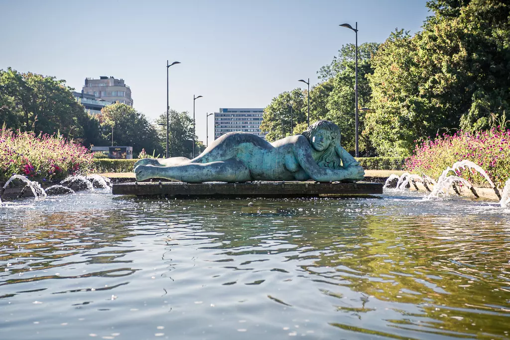 A large bronze sculpture of a reclining female figure rests in a clear pond, surrounded by colorful flowers and splashing water.