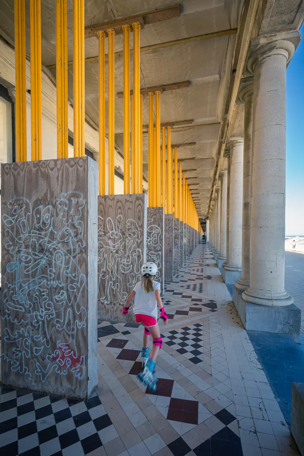 A girl in a helmet and roller skates glides down a long tiled passage with colorful columns and graffiti-covered walls.