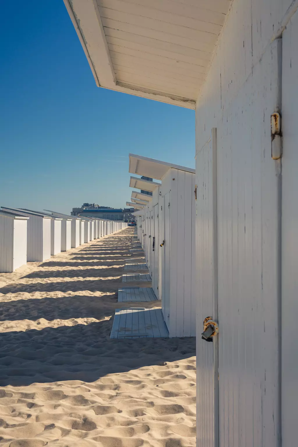 A row of white beach cabins along a sandy path, casting shadows with a clear blue sky in the background.