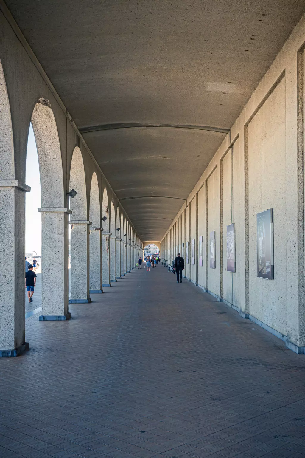 A long pedestrian walkway with arched windows, lined with columns, featuring people strolling along the paved path.