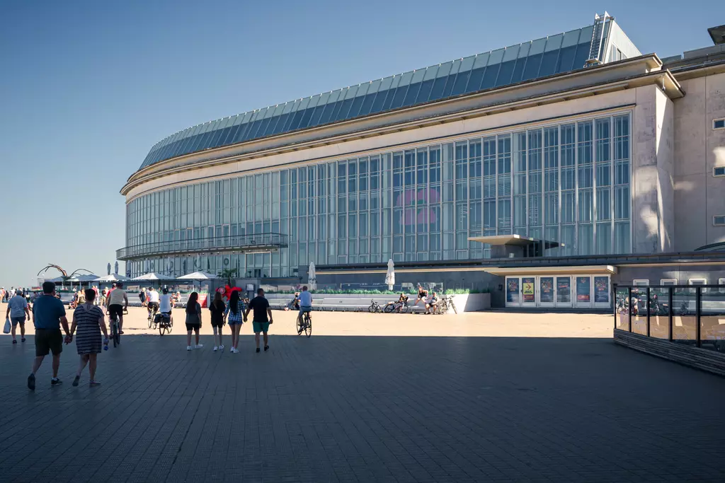 Large modern building with a glass façade, surrounded by people, bicycles, and a promenade under a clear sky.