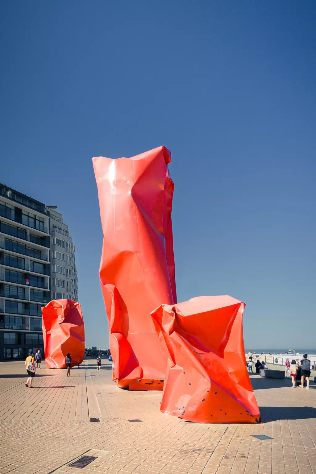 Large red sculptures with a weathered look stand in a plaza, surrounded by people and a modern building in the background.