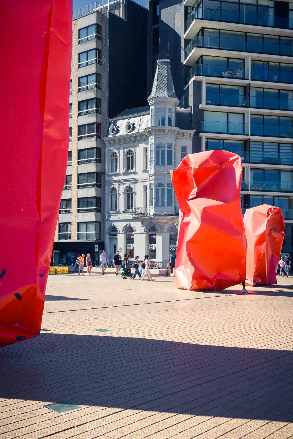 Large, crumpled red sculptures are in the foreground, with a historic building and people visible in the background.