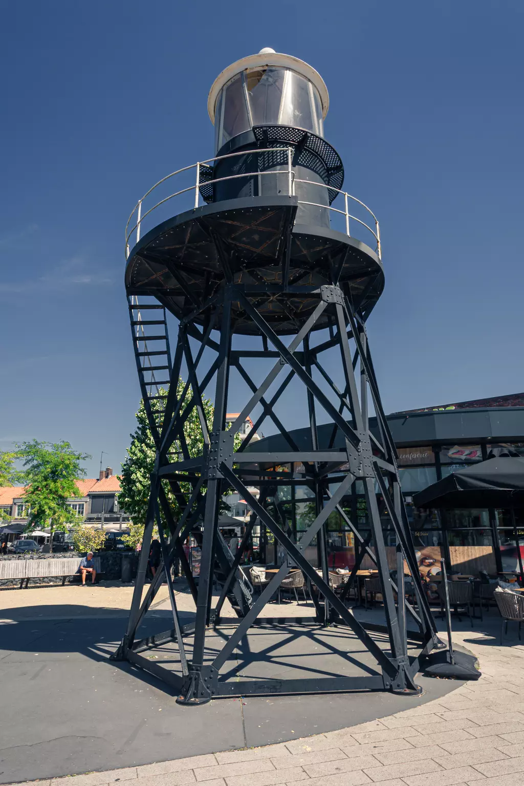 A black lighthouse with a metal staircase and transparent light cone stands in a square, surrounded by trees and cafés.