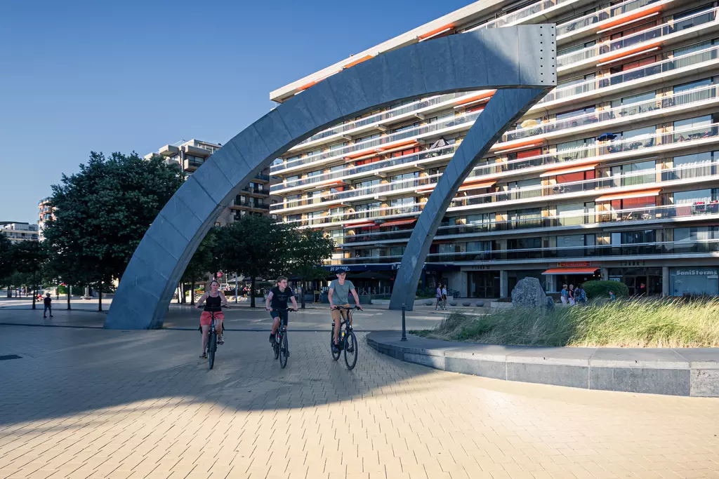 Bikers ride under a large, arching metal structure, with colorful buildings and trees in the background.