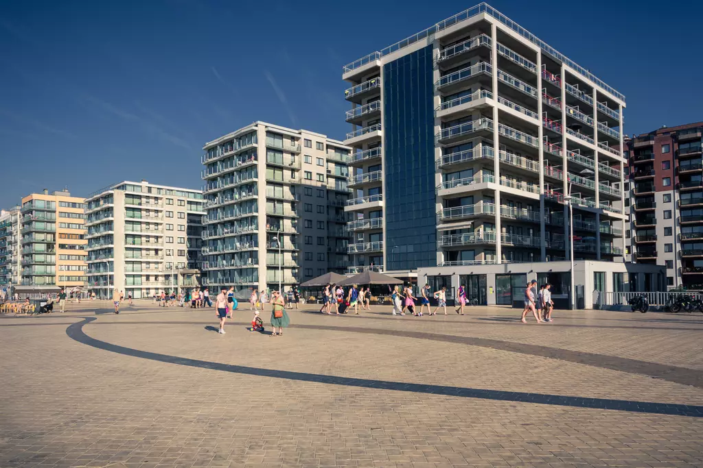 Modern buildings surround a paved area where people walk and enjoy the weather under a clear sky.